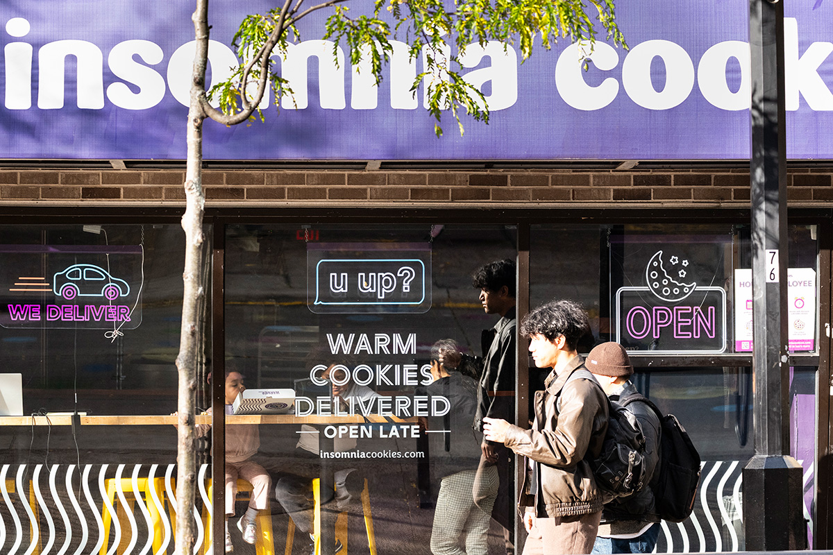 Pedestrians walk past the storefront of Insomnia Cookies, which features neon signs advertising warm cookies and late‑night delivery.
