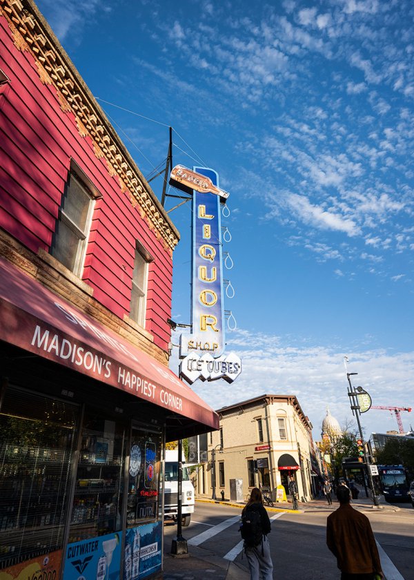 A street corner with a vintage neon liquor store sign rises above a red building under a bright blue sky in downtown Madison.