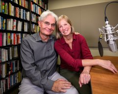 Two people sit closely together in a recording studio with bookshelves behind them and a large professional microphone positioned nearby.