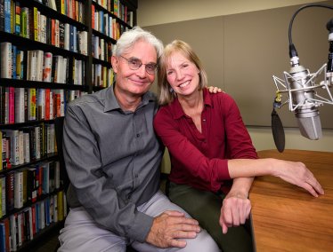 Two people sit closely together in a recording studio with bookshelves behind them and a large professional microphone positioned nearby.