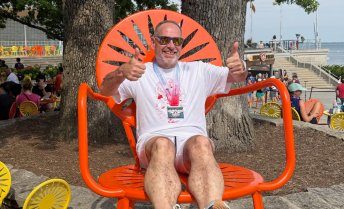 Todd Miller, wearing a white T‑shirt and marathon finisher medal, sits in a large orange terrace chair at the Memorial Union Terrace, giving two thumbs up.