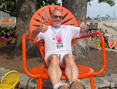 Todd Miller, wearing a white T‑shirt and marathon finisher medal, sits in a large orange terrace chair at the Memorial Union Terrace, giving two thumbs up.