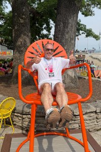 Todd Miller, wearing a white T‑shirt and marathon finisher medal, sits in a large orange terrace chair at the Memorial Union Terrace, giving two thumbs up.