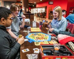 A group of people sit around a table playing a board game with cards, tokens, and a colorful game board in a lively indoor setting.