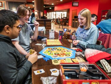 A group of people sit around a table playing a board game with cards, tokens, and a colorful game board in a lively indoor setting.
