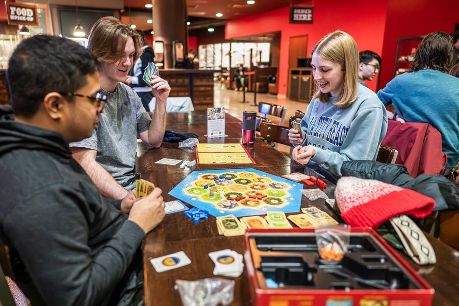 A group of people sit around a table playing a board game with cards, tokens, and a colorful game board in a lively indoor setting.