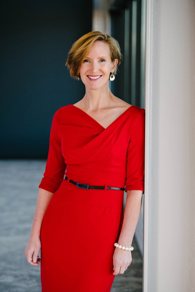 Susan Webb Yackee wearing a red dress and pearl bracelet stands leaning lightly against a wall in a modern indoor space.