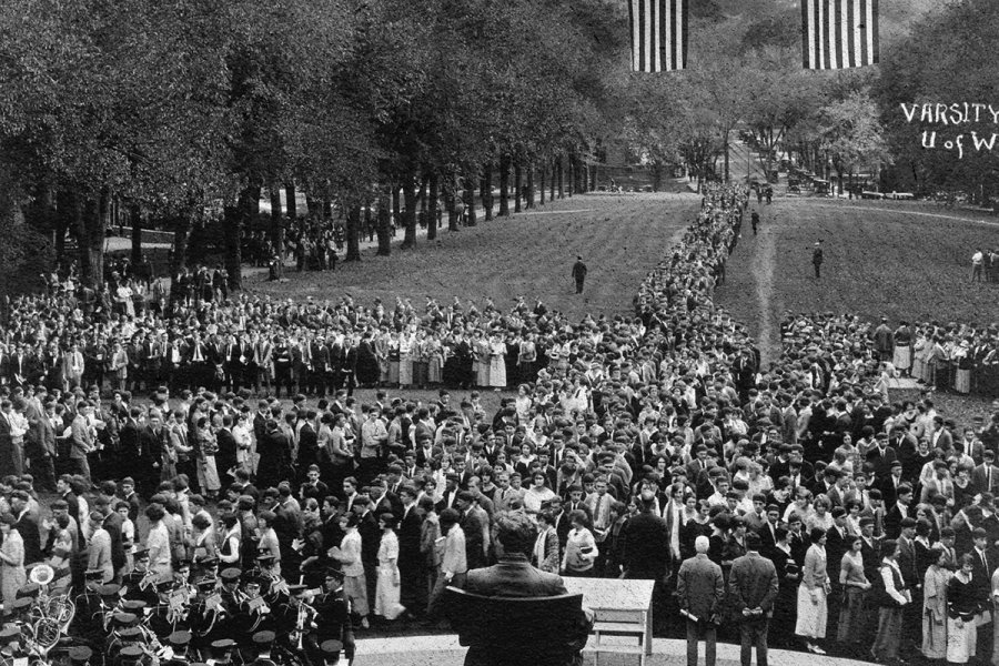 A historic black‑and‑white photograph showing a large crowd of people gathered on Bascom Hill, forming long lines that converge toward the center. Two large American flag banners hang above the scene. Trees line both sides of the lawn, and a domed capitol building is visible in the background.