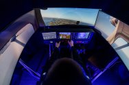 A person operates a flight simulator cockpit with hands on the controls, surrounded by illuminated instrument panels and curved screens displaying an aerial runway and landscape view.