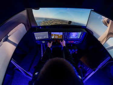 A person operates a flight simulator cockpit with hands on the controls, surrounded by illuminated instrument panels and curved screens displaying an aerial runway and landscape view.