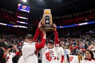 Badger women in team apparel hold an NCAA volleyball national championship trophy overhead at center court while spectators fill a large indoor arena.