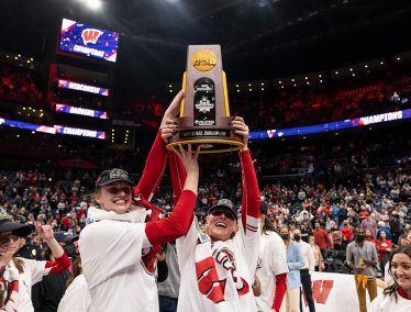 Badger women in team apparel hold an NCAA volleyball national championship trophy overhead at center court while spectators fill a large indoor arena.