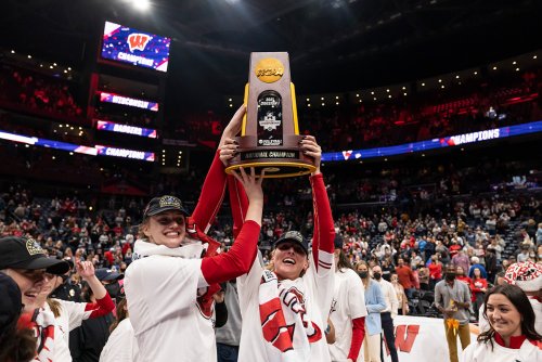 Badger women in team apparel hold an NCAA volleyball national championship trophy overhead at center court while spectators fill a large indoor arena.