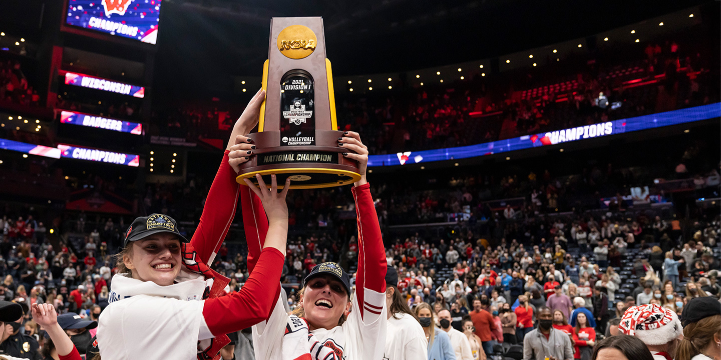 Badger women in team apparel hold an NCAA volleyball national championship trophy overhead at center court while spectators fill a large indoor arena.