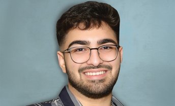 A portrait of a smiling young man with dark hair, a beard, and glasses, wearing a gray and black striped button-down shirt against a light blue textured background.