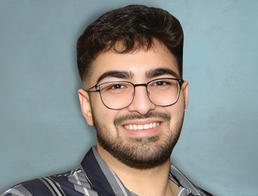 A portrait of a smiling young man with dark hair, a beard, and glasses, wearing a gray and black striped button-down shirt against a light blue textured background.