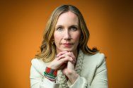 A woman with long blonde hair and colorful beaded bracelets rests her chin on her interlaced hands in a portrait against a solid orange background.
