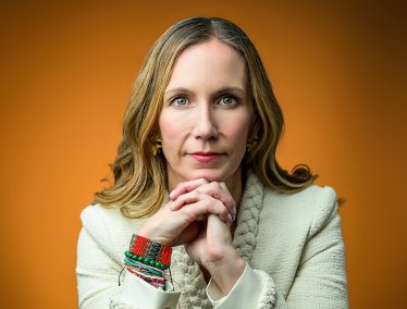 A woman with long blonde hair and colorful beaded bracelets rests her chin on her interlaced hands in a portrait against a solid orange background.
