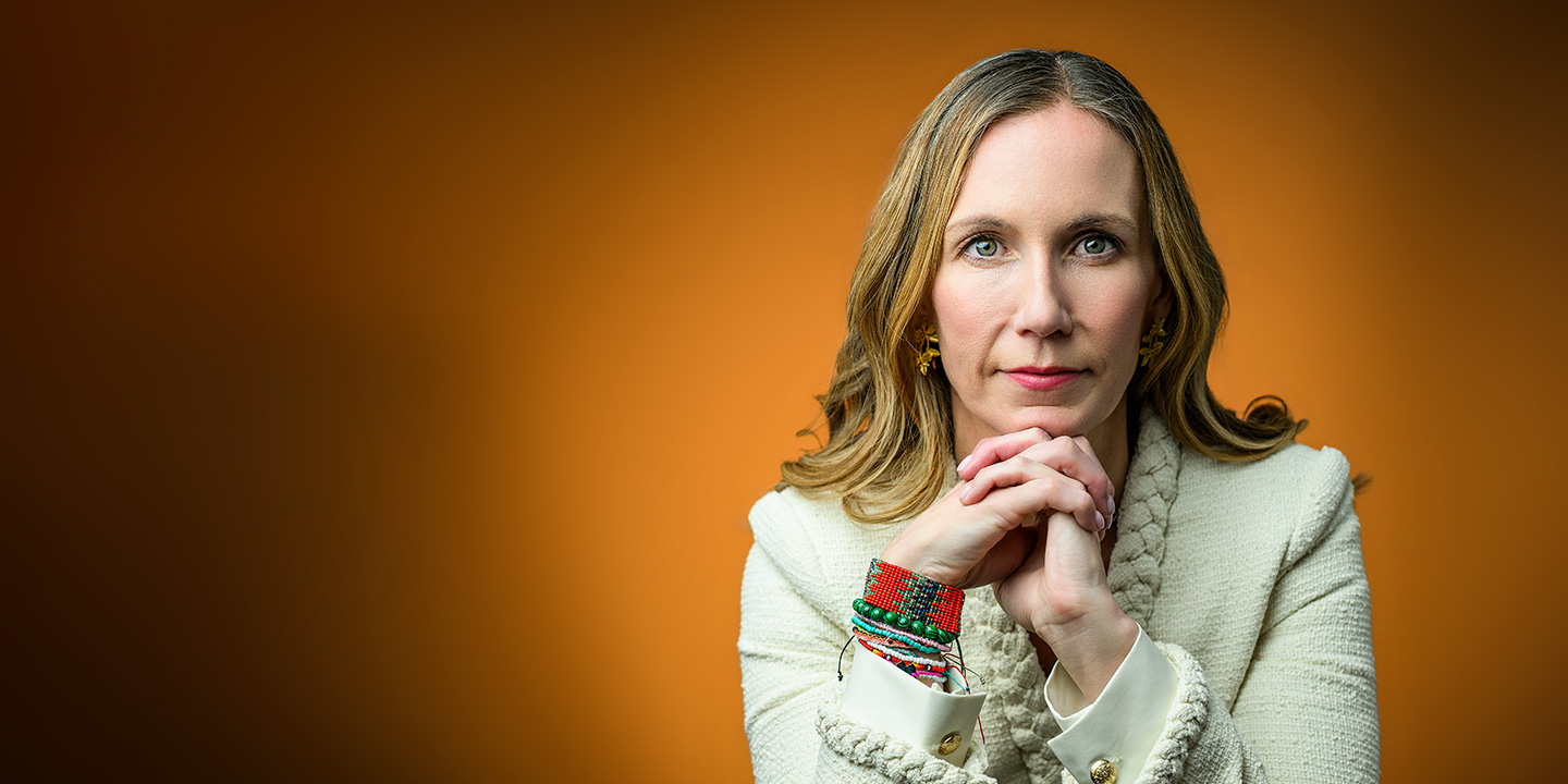 A woman with long blonde hair and colorful beaded bracelets rests her chin on her interlaced hands in a portrait against a solid orange background.