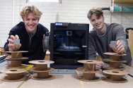 Two young men smile while holding small, square grey devices, standing behind a table with stacks of empty filament spools and a black 3D printer.