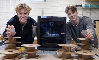 Two young men smile while holding small, square grey devices, standing behind a table with stacks of empty filament spools and a black 3D printer.