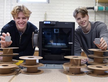 Two young men smile while holding small, square grey devices, standing behind a table with stacks of empty filament spools and a black 3D printer.