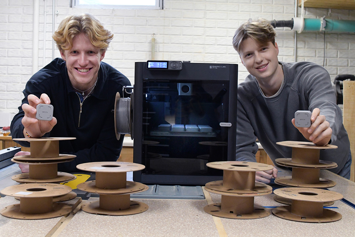 Two young men smile while holding small, square grey devices, standing behind a table with stacks of empty filament spools and a black 3D printer.