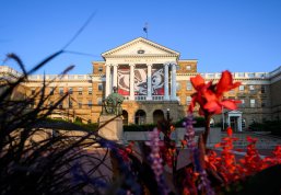 A scenic dawn view of Bascom Hall at UW–Madison, featuring a large Buckiy Badger banner behind its white pillars and the Abraham Lincoln statue in front, framed by colorful flowers in the foreground.