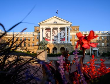 A scenic dawn view of Bascom Hall at UW–Madison, featuring a large Buckiy Badger banner behind its white pillars and the Abraham Lincoln statue in front, framed by colorful flowers in the foreground.