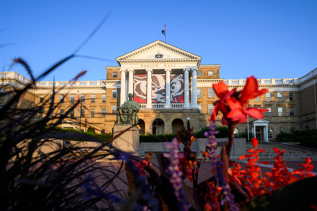 A scenic dawn view of Bascom Hall at UW–Madison, featuring a large Buckiy Badger banner behind its white pillars and the Abraham Lincoln statue in front, framed by colorful flowers in the foreground.