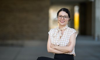A woman wearing glasses is depicted, sitting with arms crossed in an outdoor walkway, with blurred architectural columns and soft lighting in the background.
