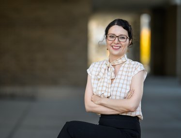 A woman wearing glasses is depicted, sitting with arms crossed in an outdoor walkway, with blurred architectural columns and soft lighting in the background.