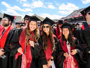 A group of smiling graduates wearing black caps and gowns with red stoles stand together in a sunny, crowded stadium during a University of Wisconsin commencement ceremony.