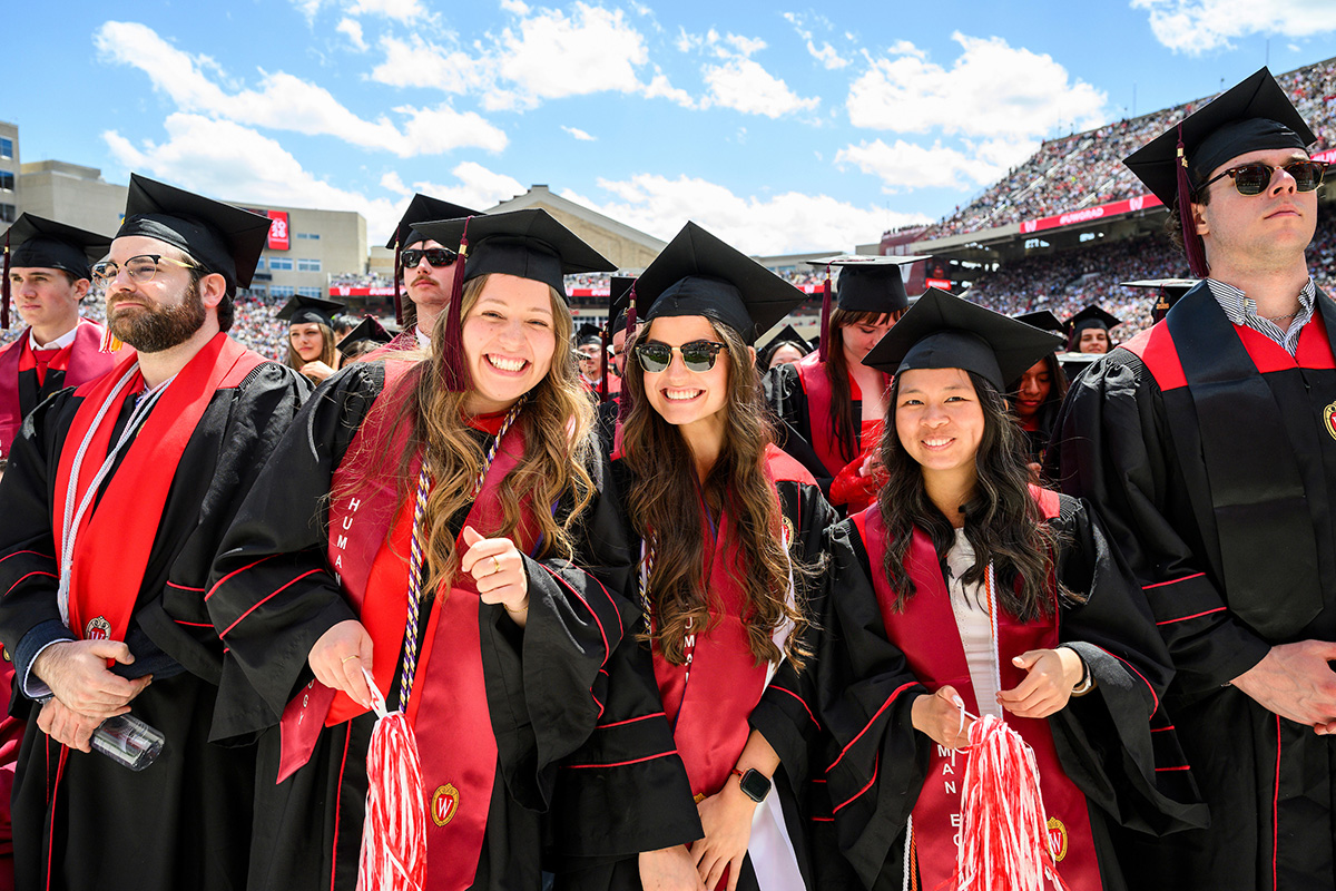 A group of smiling graduates wearing black caps and gowns with red stoles stand together in a sunny, crowded stadium during a University of Wisconsin commencement ceremony.