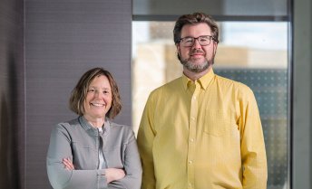 A woman and man stand together in an indoor office setting, both facing the camera, with neutral walls and a glass panel in the background.