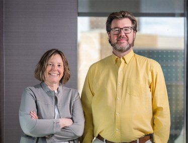 A woman and man stand together in an indoor office setting, both facing the camera, with neutral walls and a glass panel in the background.