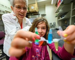 A young girl holds up a strip of plastic tubes containing colorful liquids while a woman with short grey hair watches and smiles in a laboratory setting.