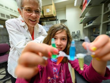 A young girl holds up a strip of plastic tubes containing colorful liquids while a woman with short grey hair watches and smiles in a laboratory setting.