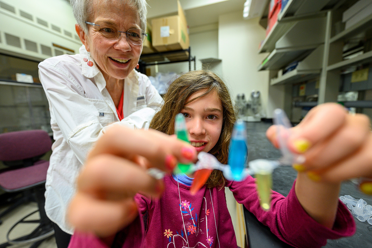 A young girl holds up a strip of plastic tubes containing colorful liquids while a woman with short grey hair watches and smiles in a laboratory setting.