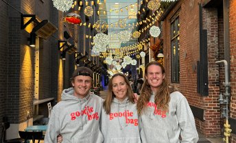 Three people stand arm-in-arm smiling in an outdoor alleyway decorated with hanging globe lights, each wearing a grey hoodie with "goodie bag" printed in red lettering across the chest.