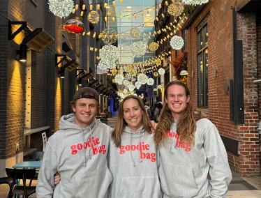 Three people stand arm-in-arm smiling in an outdoor alleyway decorated with hanging globe lights, each wearing a grey hoodie with "goodie bag" printed in red lettering across the chest.