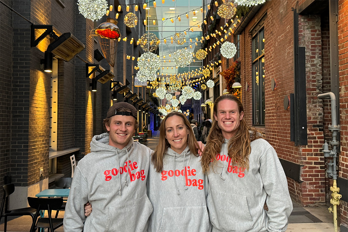 Three people stand arm-in-arm smiling in an outdoor alleyway decorated with hanging globe lights, each wearing a grey hoodie with "goodie bag" printed in red lettering across the chest.