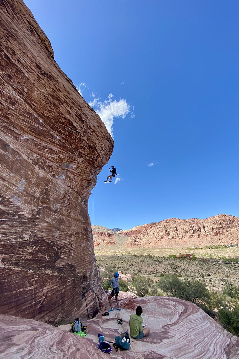 A rock climber rappels from a massive red sandstone overhang while two others watch from a flat rocky ledge below against a clear blue sky and desert mountain landscape.