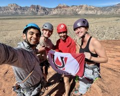 Four rock climbers wearing helmets and climbing harnesses smile for a selfie while holding a 'Wisconsin Hoofers' flag on a sandstone peak with a desert mountain range in the background.