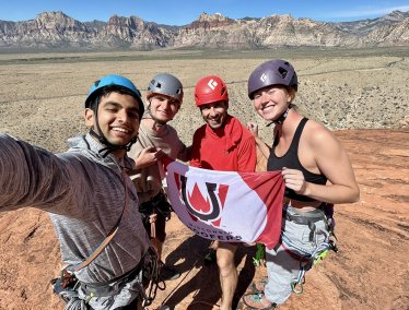 Four rock climbers wearing helmets and climbing harnesses smile for a selfie while holding a 'Wisconsin Hoofers' flag on a sandstone peak with a desert mountain range in the background.