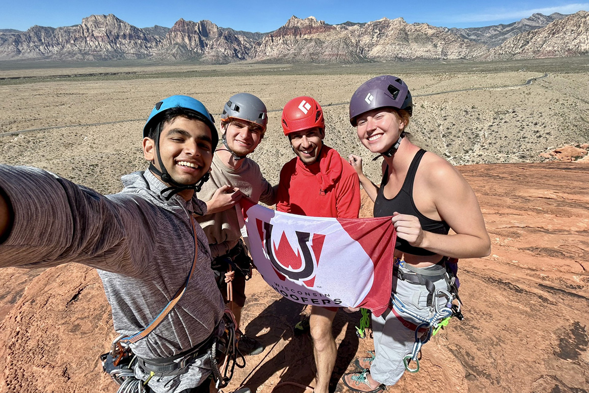 Four rock climbers wearing helmets and climbing harnesses smile for a selfie while holding a 'Wisconsin Hoofers' flag on a sandstone peak with a desert mountain range in the background.
