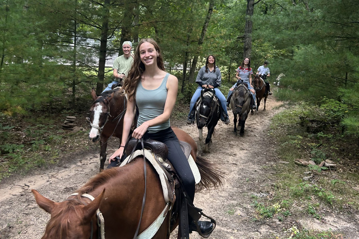 A group of five people ride horses in a single-file line down a dirt trail through a lush green forest, with the lead rider smiling at the camera.