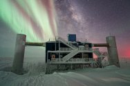 A snow-covered research facility in Antarctica stands under a starry night sky illuminated by the vibrant green and red ribbons of the aurora australis and the Milky Way galaxy.