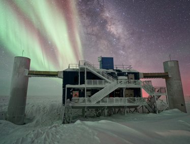 A snow-covered research facility in Antarctica stands under a starry night sky illuminated by the vibrant green and red ribbons of the aurora australis and the Milky Way galaxy.