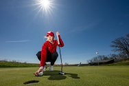 A female golfer in a red jacket and University of Wisconsin cap crouches on a green to place a golf ball near a hole while holding a putter under a clear blue sky.